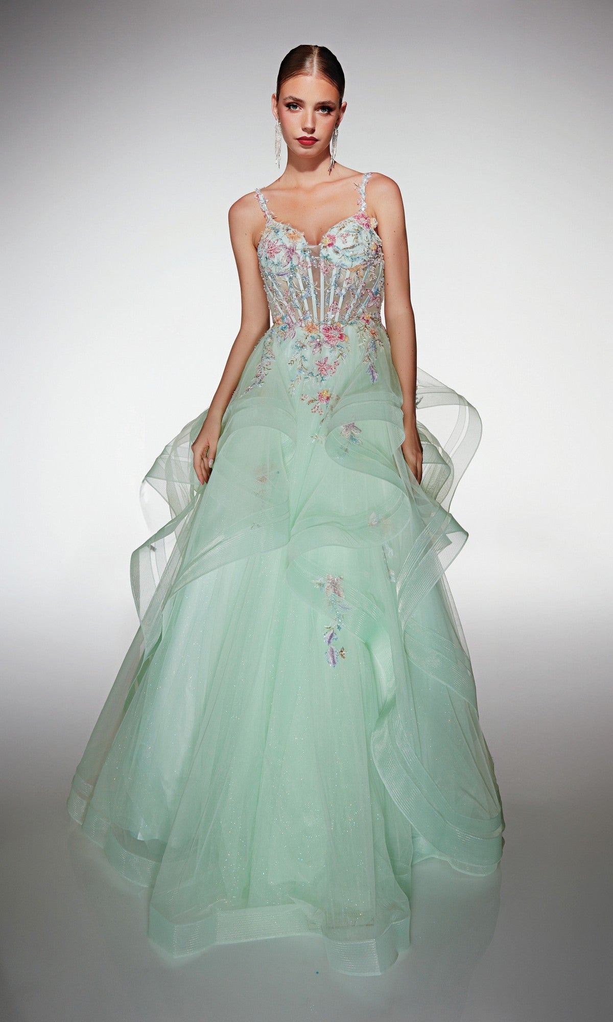 A woman wears an ice blue Alyce 62201 long formal dress featuring 3D floral appliques, a sheer bodice, and a layered tulle skirt, standing against a plain light background.
